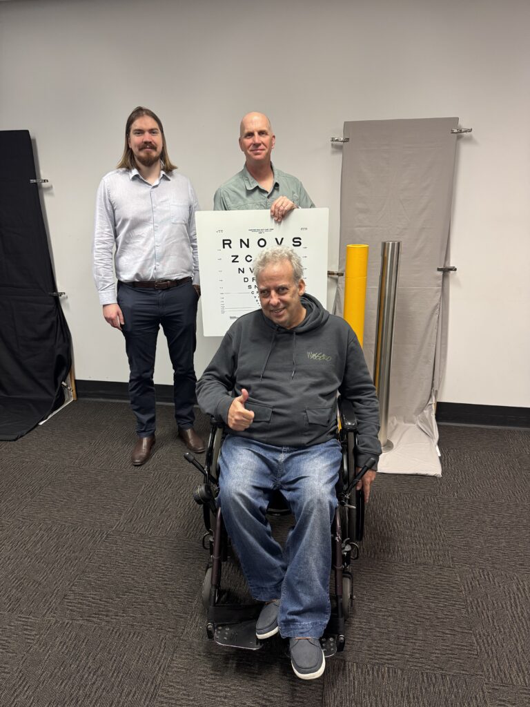 A man in a wheelchair wearing blue jeans and a grey jumper giving a “thumbs up”. He is in front of 2 other men and an eye chart