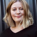 Bec has short blonde hair and wears a black v neck t-shirt.