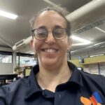 Kristy is wearing a navy-blue polo shirt, with her hair tied up, and glasses.