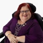 Karen is wearing a dark purple shirt with a long silver necklace. She has glasses on with shoulder length hair. Karen is sitting in her wheelchair and smiling at the camera.