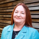 Teresa has long red hair and is wearing a pale blue blazer with a black shirt.