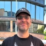 Alan has glasses on and wears a black cap and a black t-shirt.