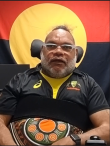 A man using a wheelchair wearing glasses, he also has an Indigenous flag behind him and a black shirt.