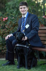 A man in a blue suit with a dark blue tie sitting in a garden with a guide dog