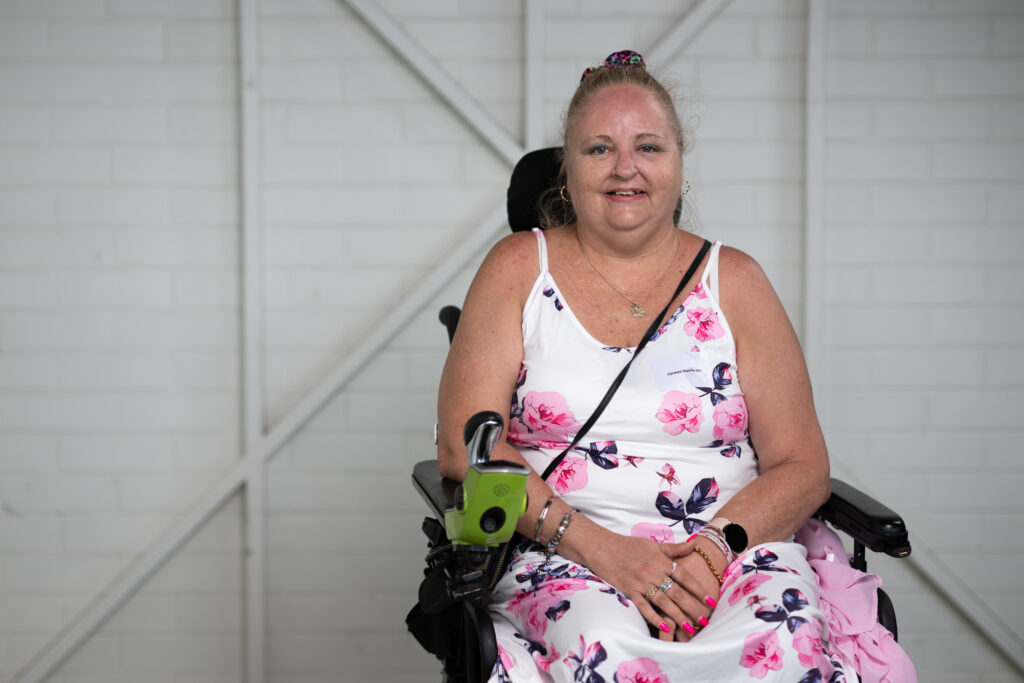 Carmen is smiling for the photo, sitting in her electric wheelchair. She has blonde hair tied back in a high bun. She wears a white and pink floral dress.