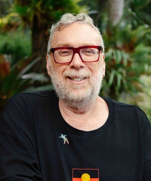 Man in a pair of red rimmed glasses and a black shirt that has an indigenous flag. He is standing in a garden.