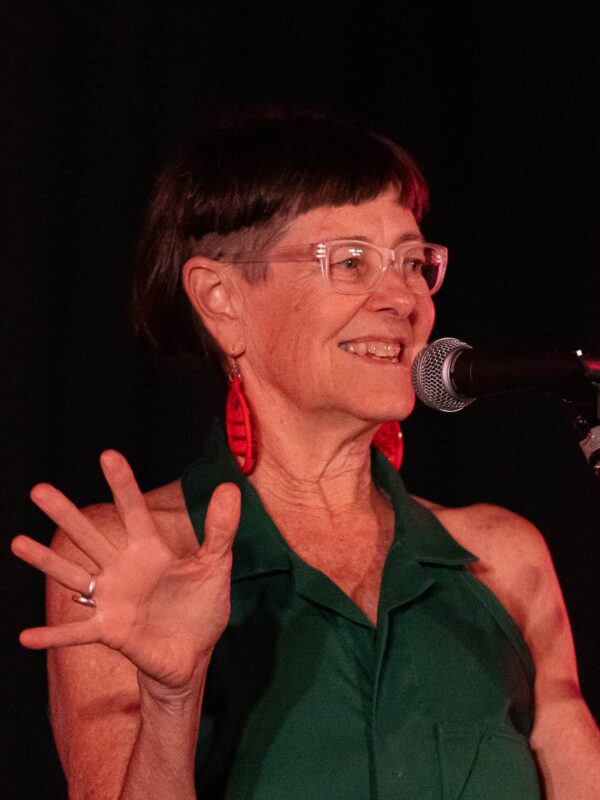 Woman with very short brown hair and is wearing a dark green coloured blouse with red earrings and clear glasses.