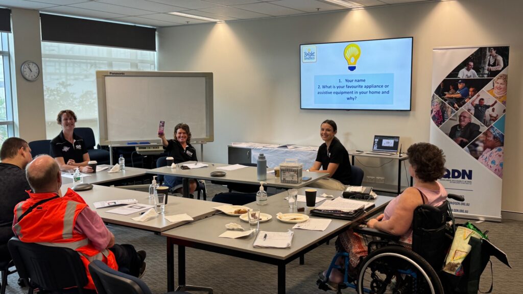2 Energy Champions, 3 workshop attendees and a QDN staff member sitting around a central desk in a room. There is a screen projecting Bright Sparks content . The attendees have Bright Sparks material on their desks.