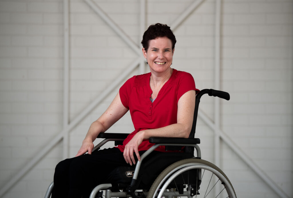Kate is wearing a red shirt, while smiling at the camera. She is in a wheelchair, with dark short brown hair.