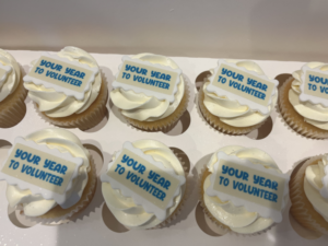 A tray of cupcakes with white icing and a blue worded sign on top saying Your year to volunteer