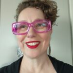 A woman with short curly hair, pink framed glasses and a black top smiling at the camera.