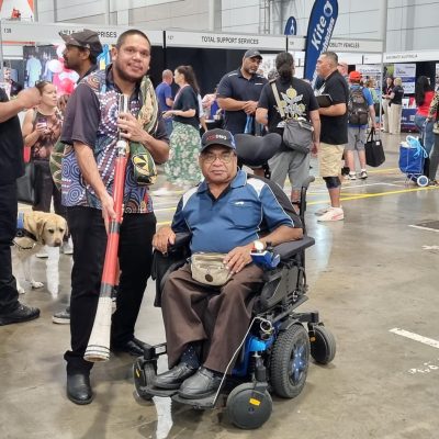 A photo of two people at the Brisbane Disability Connection. One person uses a wheelchair, the other person is standing, holding an Aboriginal musical instrument in is hands. Both people are looking at the camera smiling.