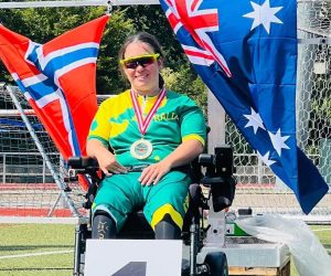 Amy Tobin sitting in a power wheelchair, sitting on the first-place podium, wearing a green-and-gold Australia uniform, sunglasses, and a medal, with Australian flags behind on an athletics field.