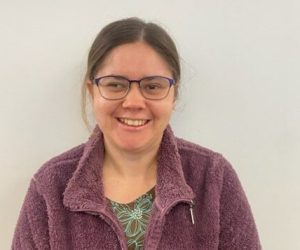 Danielle is smiling for the camera. Her brown hair is tied back, she wears glasses and purple zip up jumper.