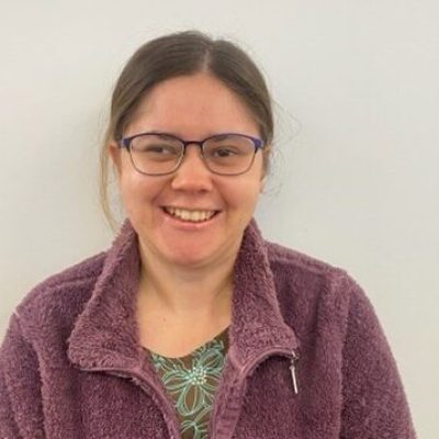 Danielle is smiling for the camera. Her brown hair is tied back, she wears glasses and purple zip up jumper.