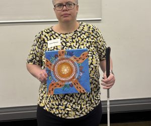 Daisy is wearing a black and cream top and black pants. She is holding up her artwork in one hand and holds her white cane in the other.