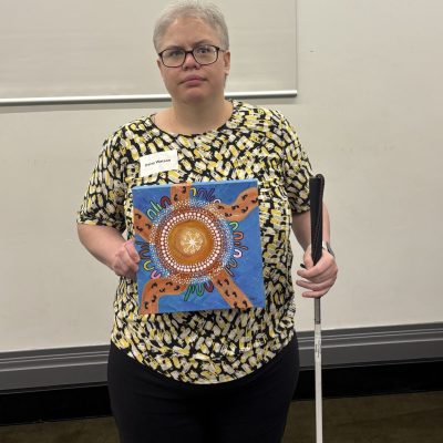 Daisy is wearing a black and cream top and black pants. She is holding up her artwork in one hand and holds her white cane in the other.