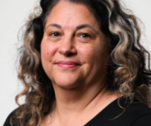 Female with long brown curly hair, and a black top, smiling at the camera