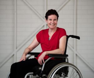 Kate is wearing a red shirt, while smiling at the camera. She is in a wheelchair, with dark short brown hair.