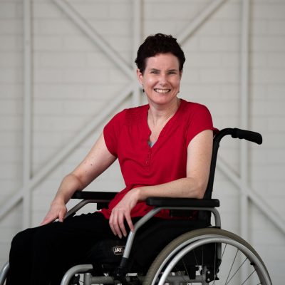Kate is wearing a red shirt, while smiling at the camera. She is in a wheelchair, with dark short brown hair.