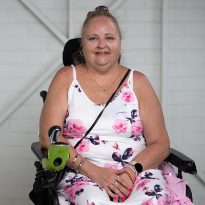 Carmen is smiling for the photo, sitting in her electric wheelchair. She has blonde hair tied back in a high bun. She wears a white and pink floral dress.